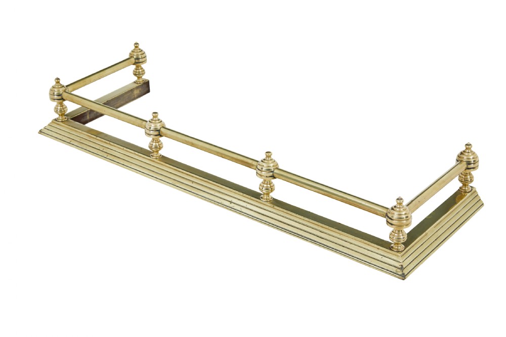 An early 20th-century polished brass fireside fender featuring a stepped base and a raised top rail supported by turned spherical finials.