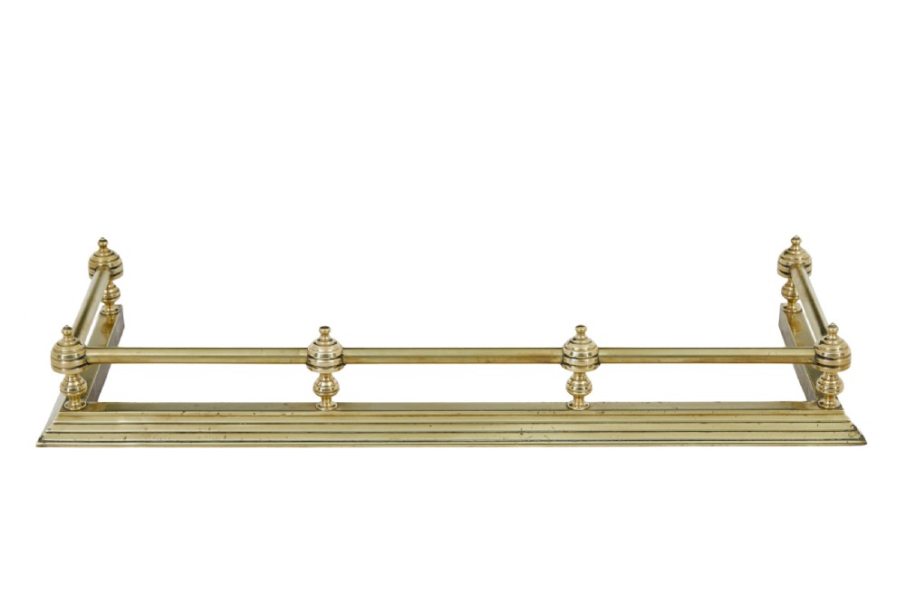 An early 20th-century polished brass fireside fender featuring a stepped base and a raised top rail supported by turned spherical finials. View II