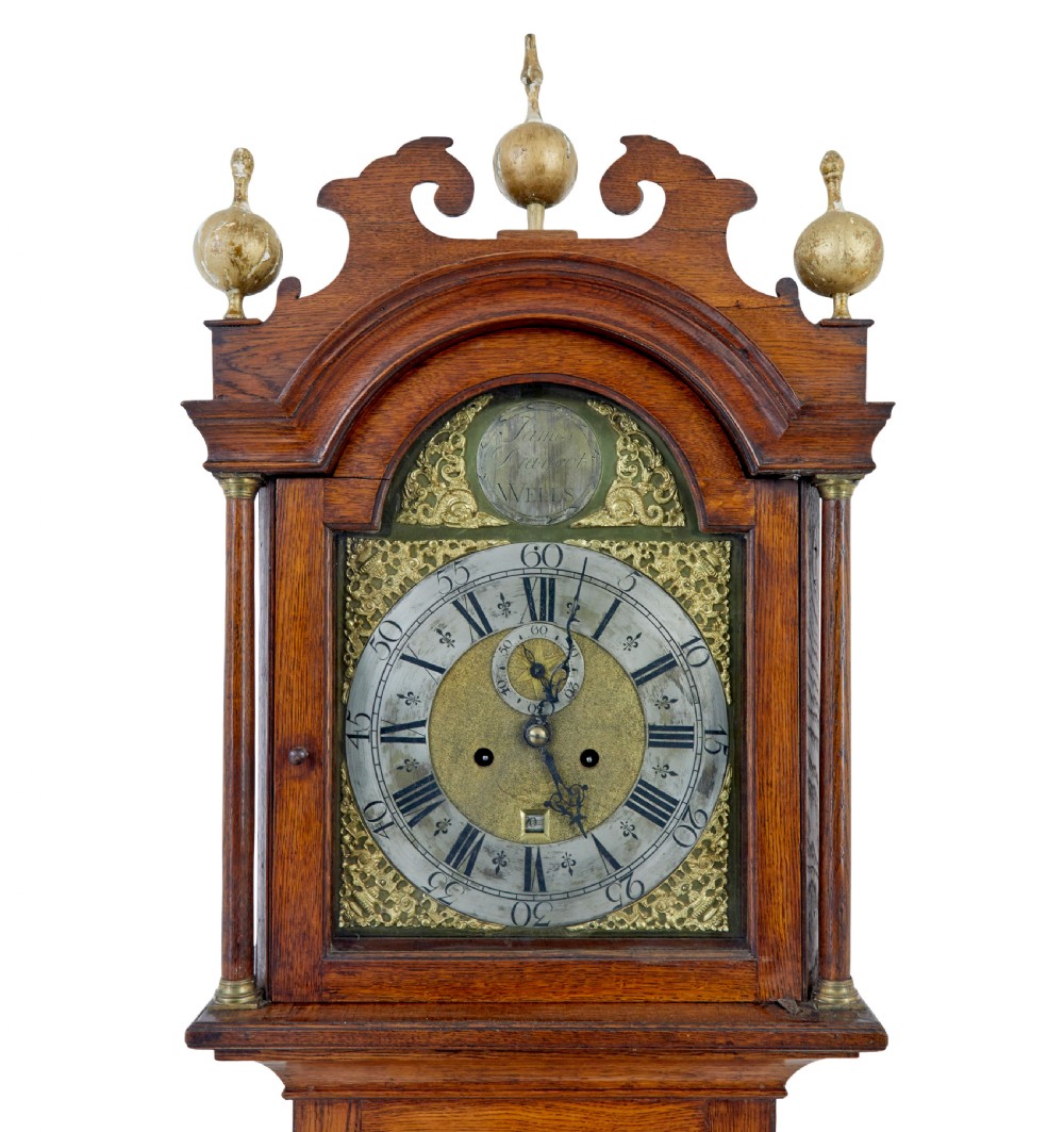 An 18th-century oak longcase clock featuring a brass dial, pediment hood with brass finials, rectangular trunk door, and plinth base. View VI