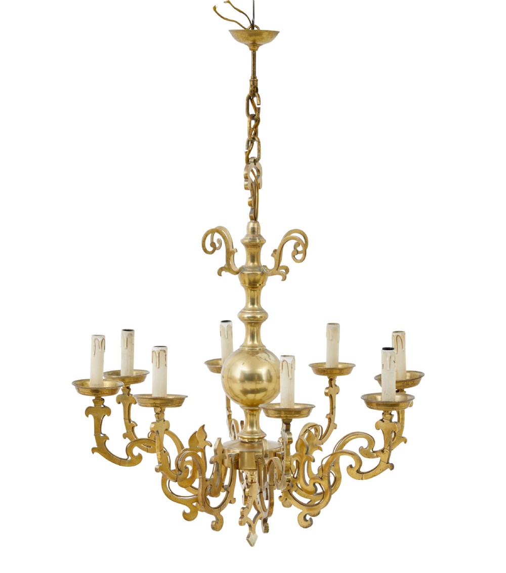 An early 20th century brass eight-arm chandelier featuring scrolling branches, circular drip pans, candle sleeves, a central baluster stem, and a suspension chain.