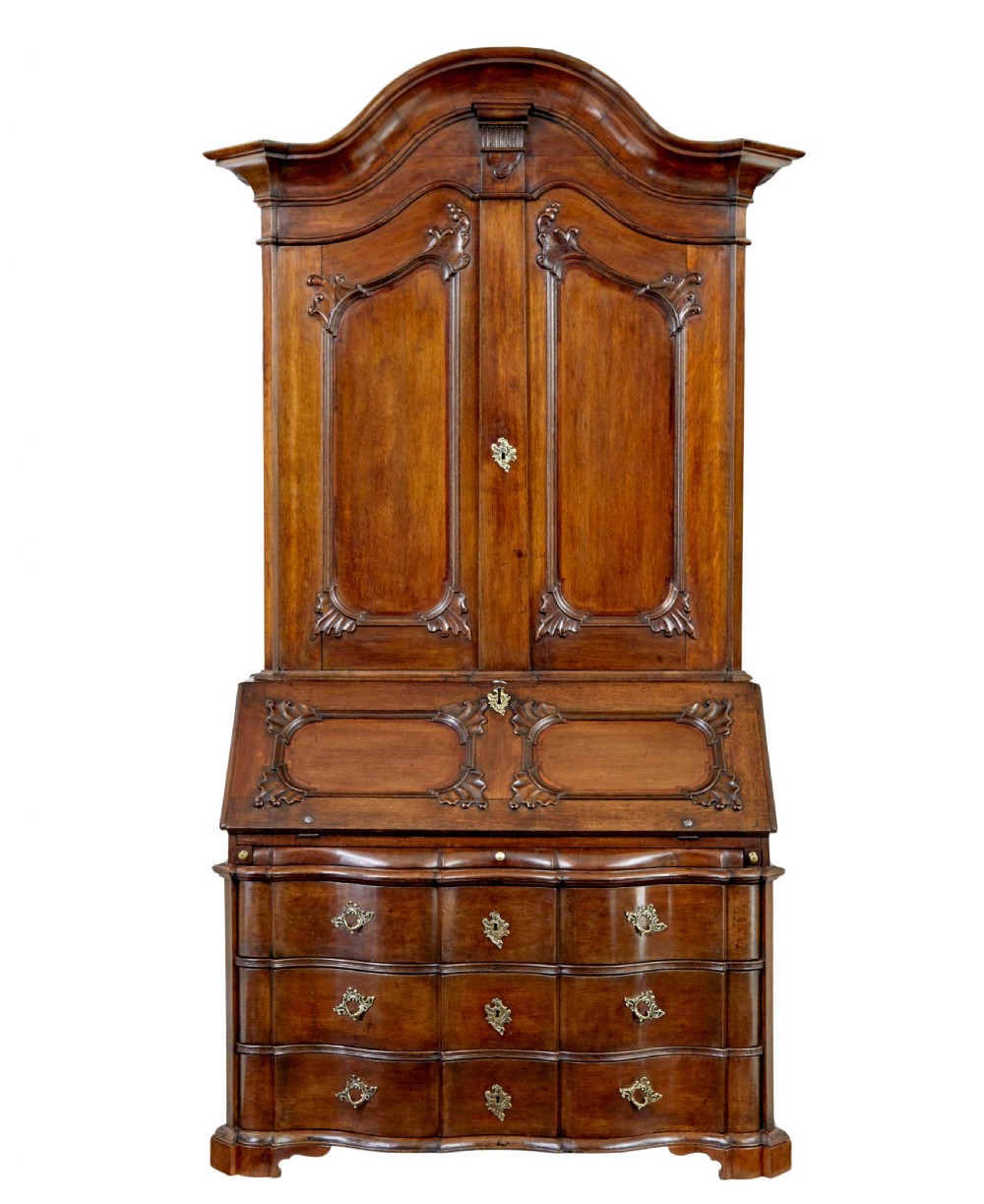An 18th-century Norwegian carved oak bureau bookcase featuring an arched pediment, panelled upper doors, a slant-front writing slant, serpentine lower drawers, and brass escutcheons.