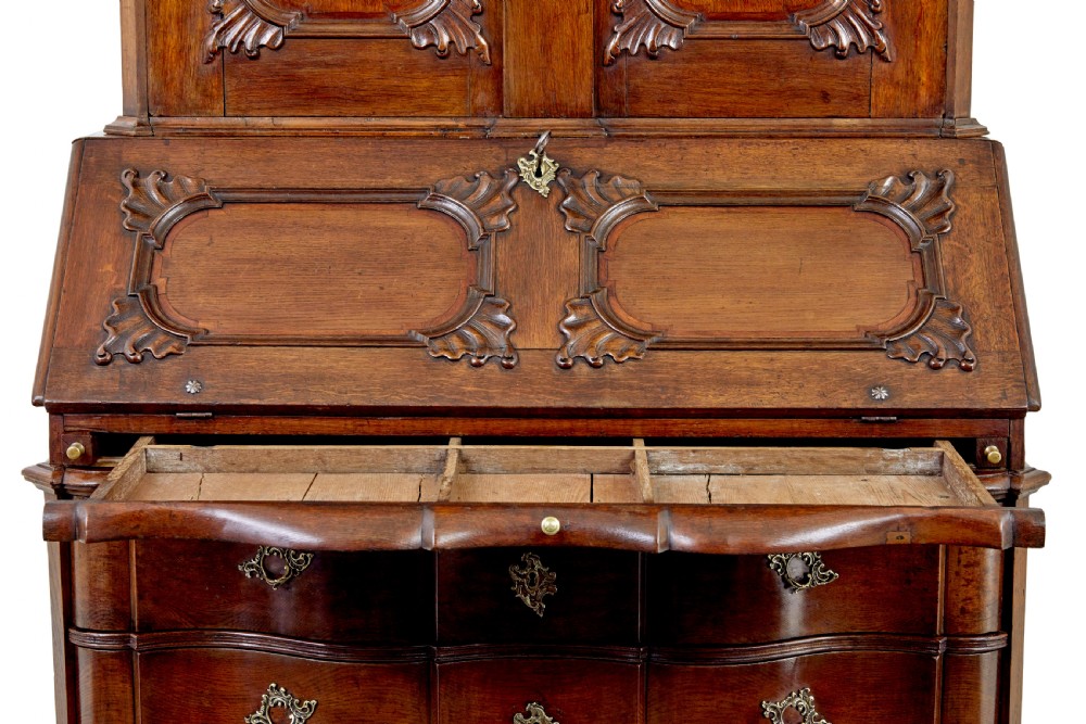 An 18th-century Norwegian carved oak bureau bookcase featuring an arched pediment, panelled upper doors, a slant-front writing slant, serpentine lower drawers, and brass escutcheons. View X