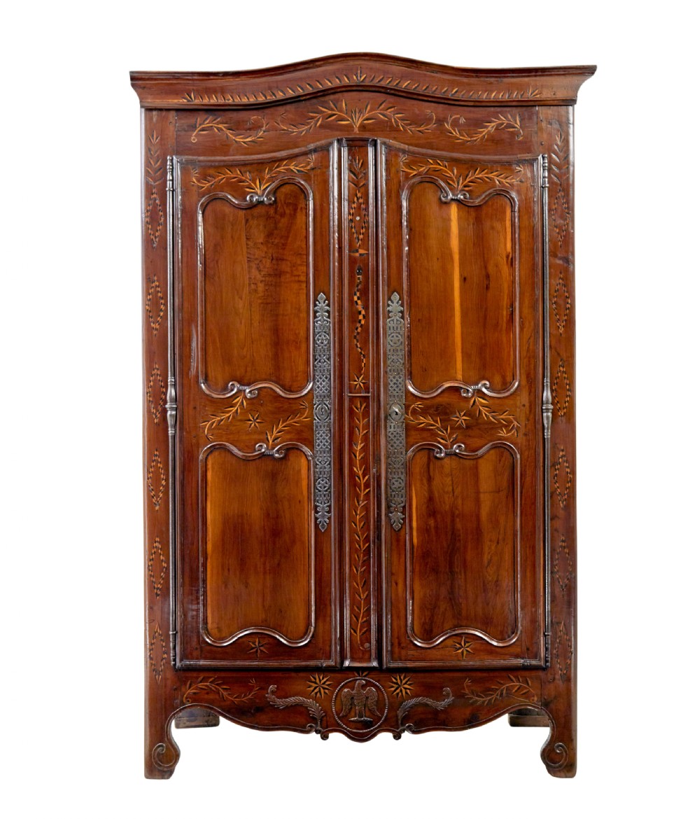 An 18th-century French armoire in warm brown yew and chestnut wood featuring carved foliate inlay, arched panel doors, and long iron escutcheons.