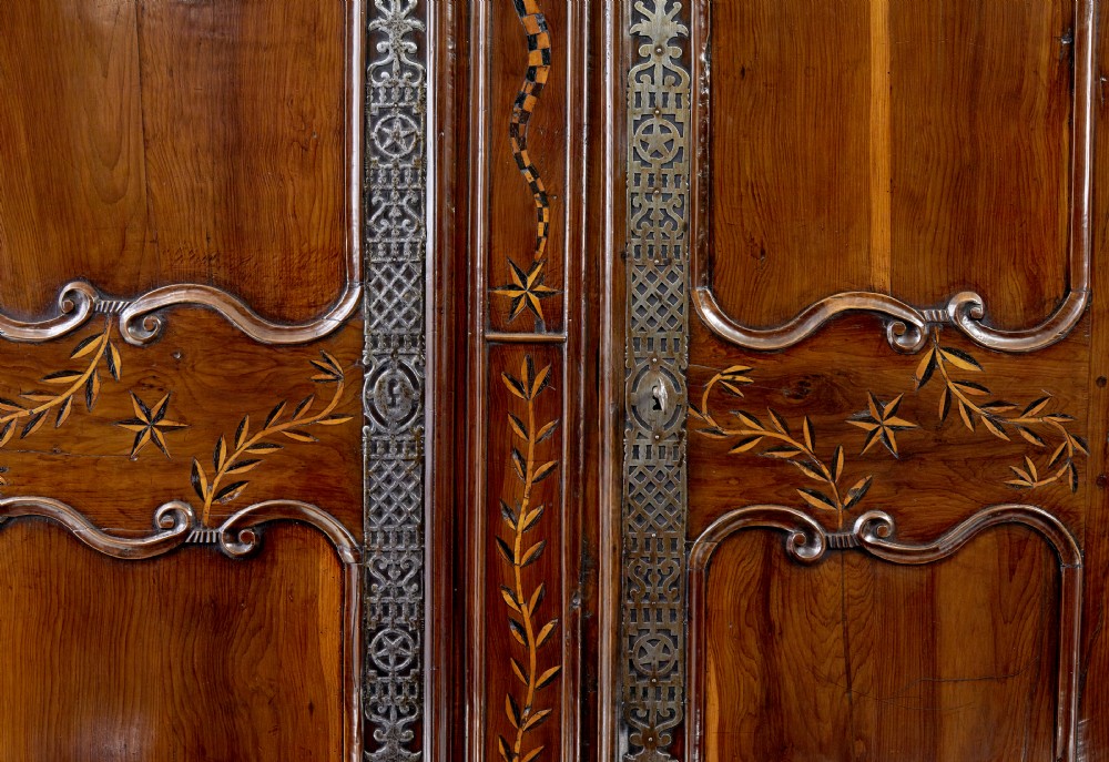 An 18th-century French armoire in warm brown yew and chestnut wood featuring carved foliate inlay, arched panel doors, and long iron escutcheons. View V