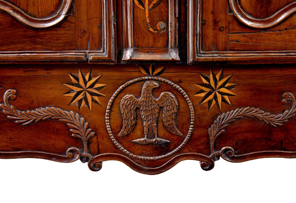 An 18th-century French armoire in warm brown yew and chestnut wood featuring carved foliate inlay, arched panel doors, and long iron escutcheons. View IX