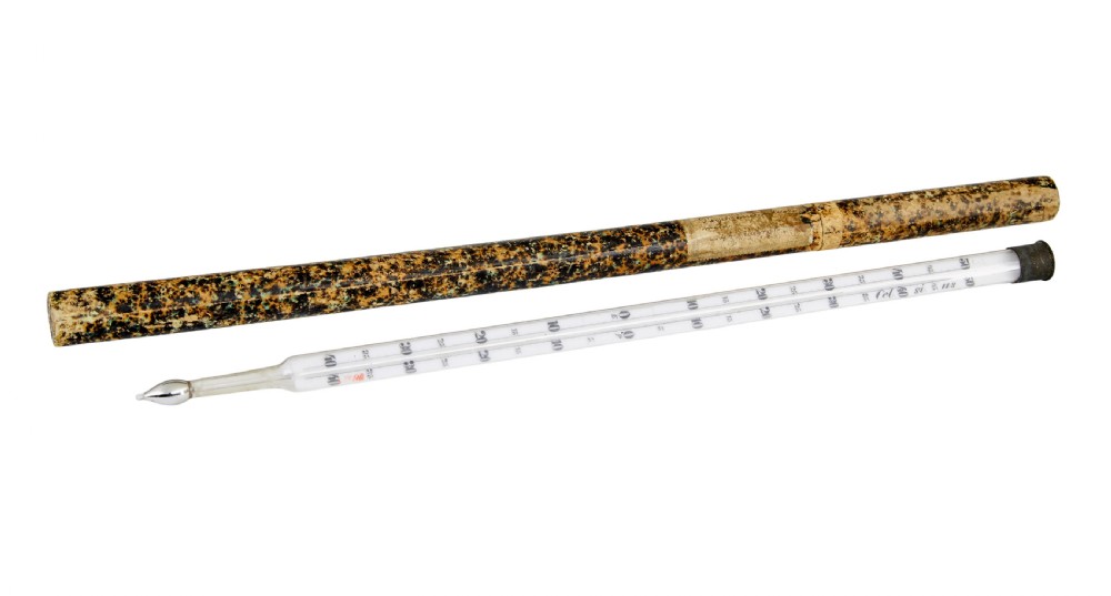A 19th-century clear glass thermometer with printed measurement scales resting alongside its original brown marbled cylindrical paperboard storage tube.
