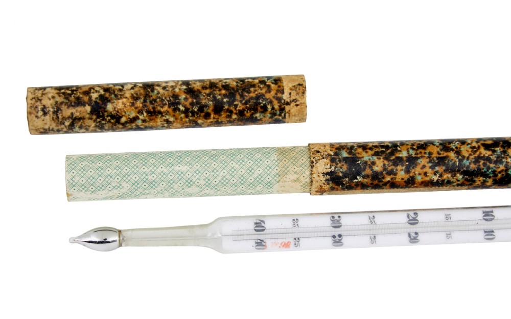 A 19th-century clear glass thermometer with printed measurement scales resting alongside its original brown marbled cylindrical paperboard storage tube. View III