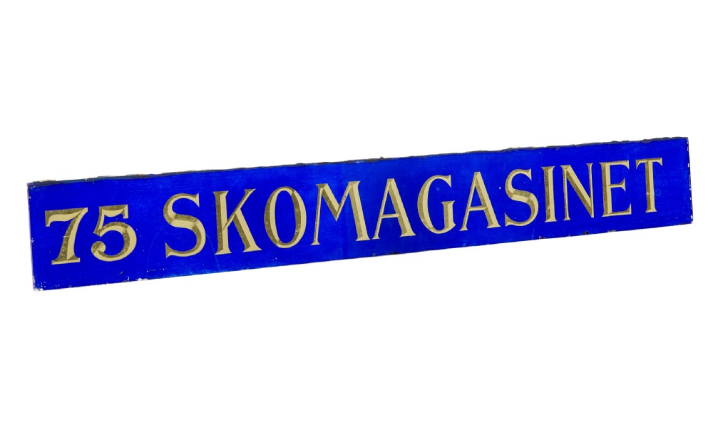 A long rectangular cobalt blue glass sign with gilded gold leaf lettering reading 75 SKOMAGASINET.