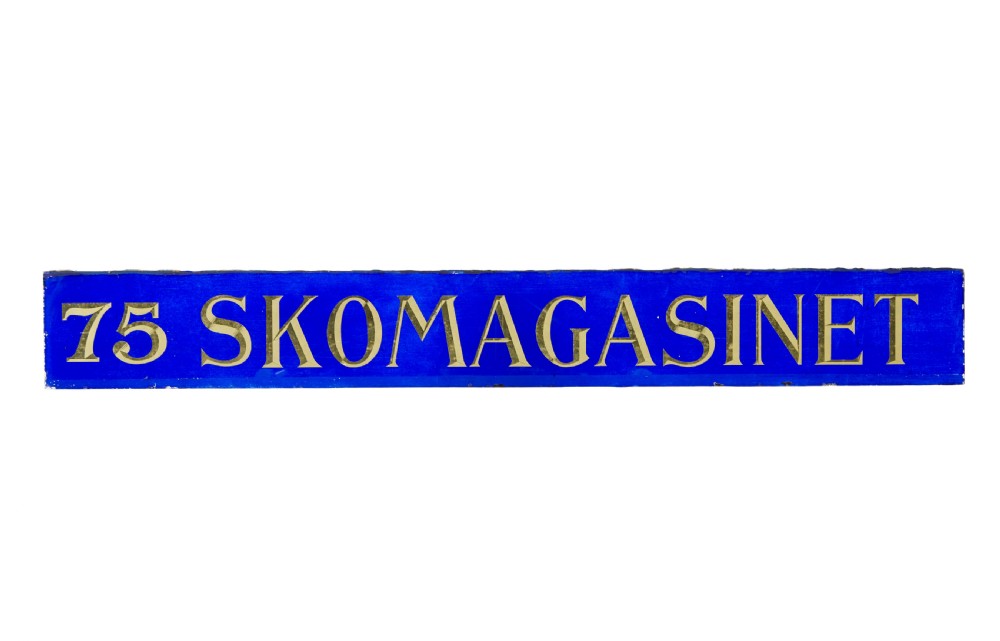 A long rectangular cobalt blue glass sign with gilded gold leaf lettering reading 75 SKOMAGASINET. View II