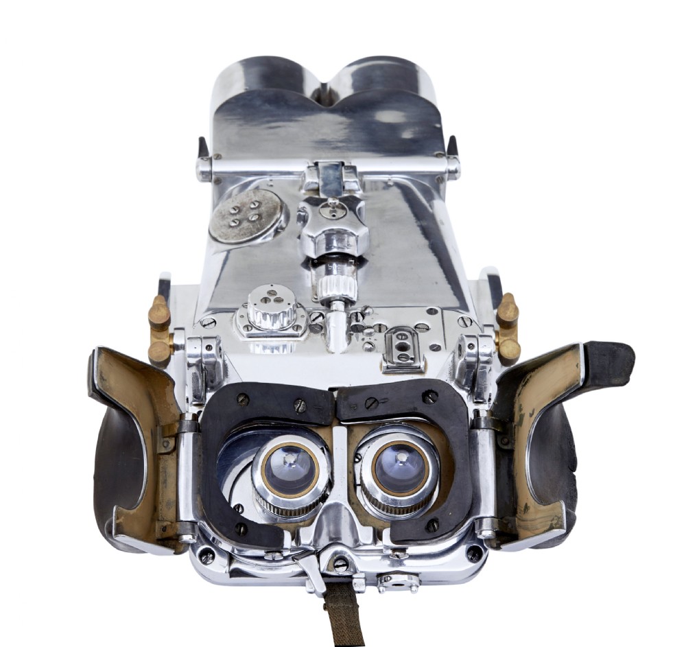 A pair of polished steel military binoculars mounted on a tall three-legged metal tripod stand against a plain background. View V