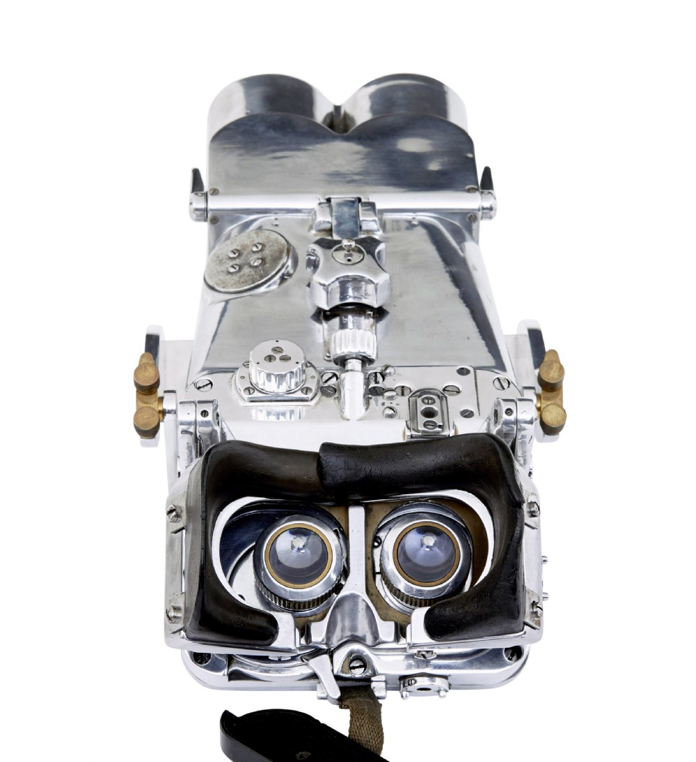 A pair of polished steel military binoculars mounted on a tall three-legged metal tripod stand against a plain background. View VI