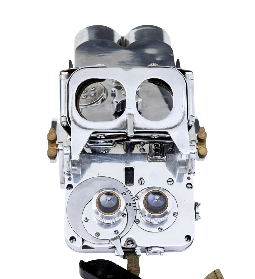 A pair of polished steel military binoculars mounted on a tall three-legged metal tripod stand against a plain background. View VII