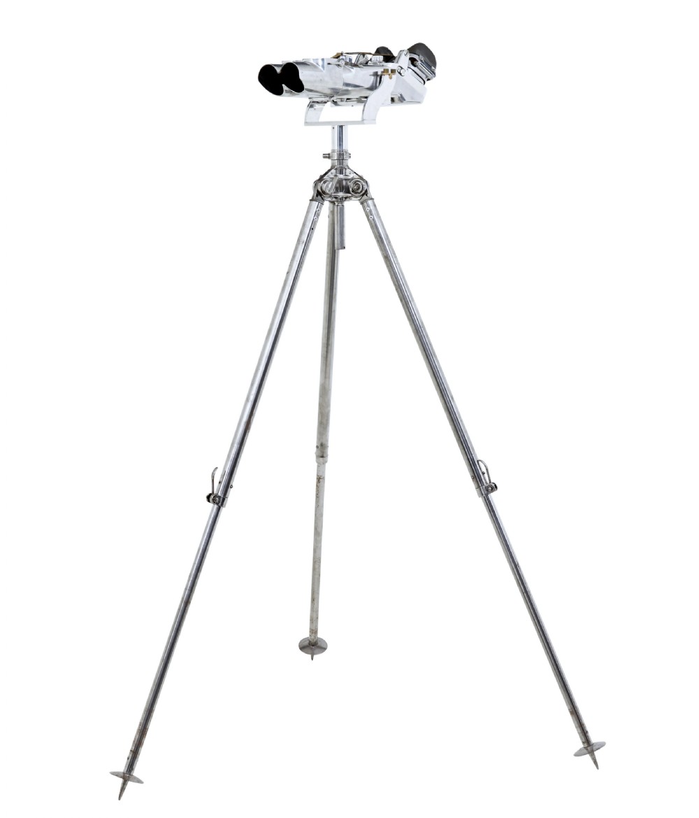 A pair of polished steel military binoculars mounted on a tall three-legged metal tripod stand against a plain background. View XI