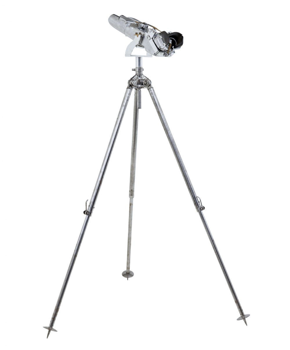 A pair of polished steel military binoculars mounted on a tall three-legged metal tripod stand against a plain background. View XII