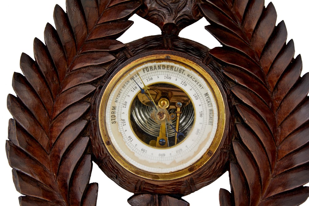 A 19th-century dark carved walnut aneroid wall barometer featuring foliate leaf carvings surrounding a central thermometer tube and a lower circular glass dial. View II