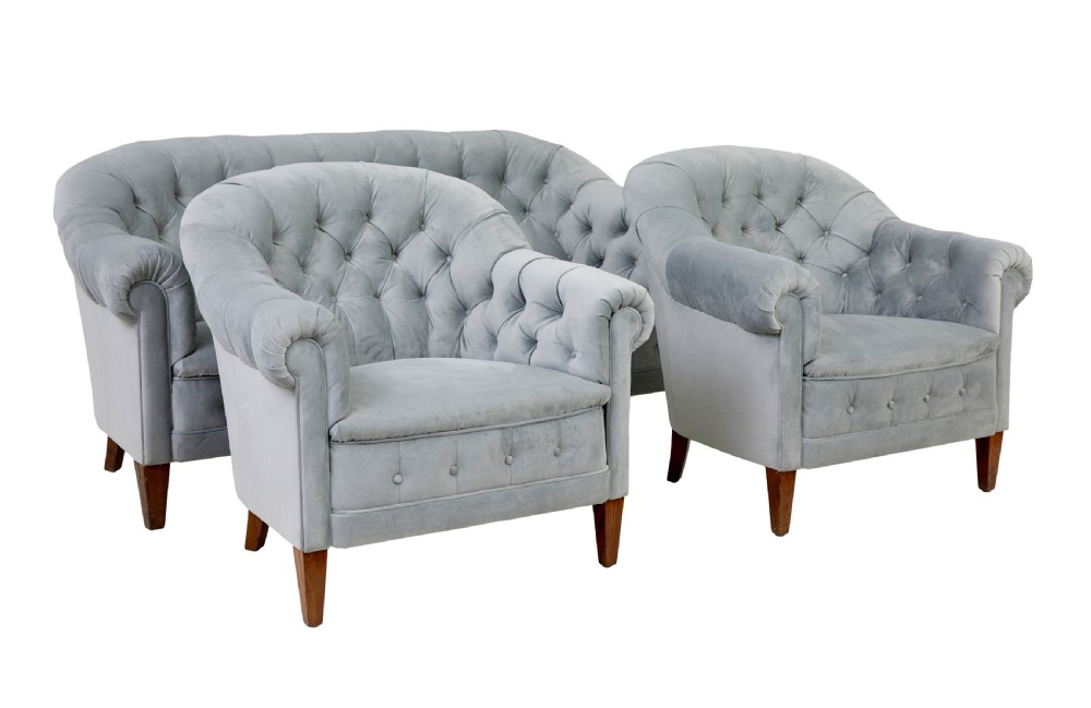 An early 20th-century three-piece suite comprising a two-seater sofa and two matching armchairs upholstered in light grey deep-buttoned velvet with tapered wooden legs.
