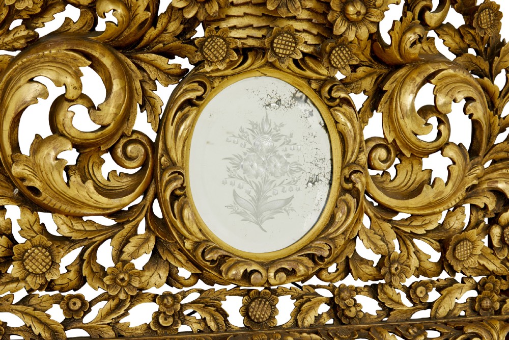 An ornate gold gilt wall mirror featuring a carved cushion frame decorated with floral motifs, scrollwork, and an etched oval glass crest. View II