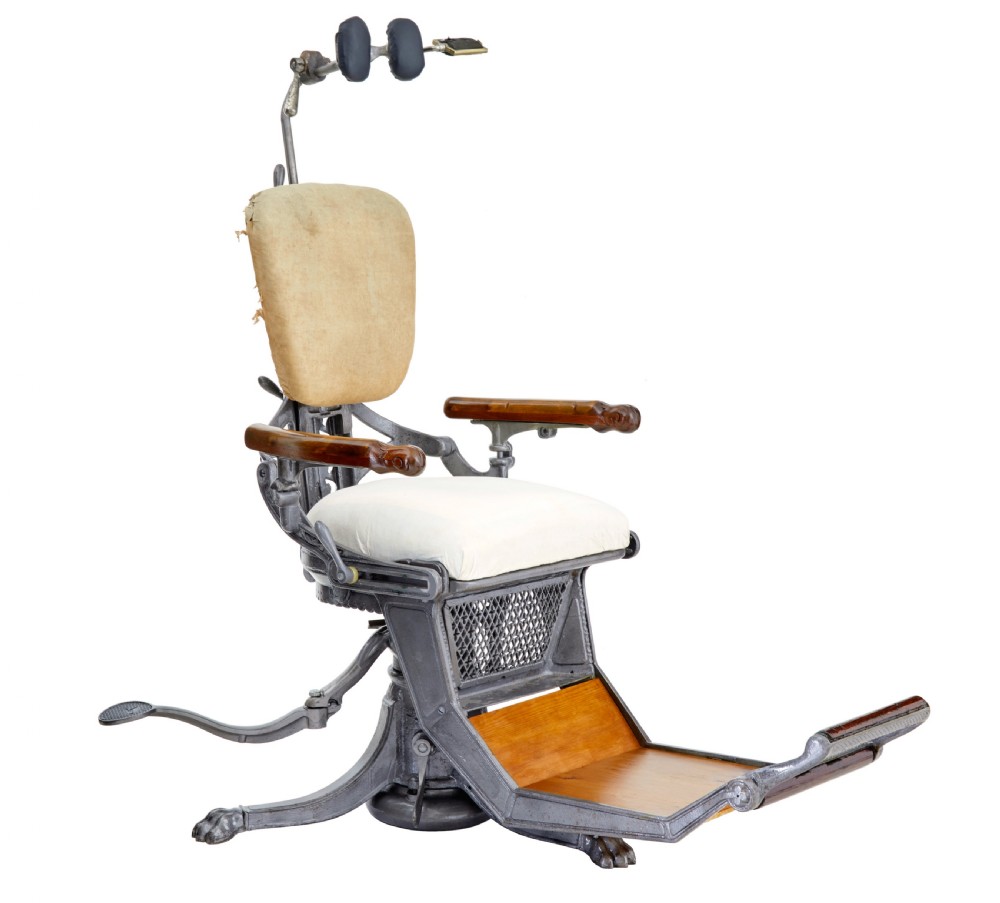 A 19th-century American cast iron dentist chair featuring an adjustable headrest, cream upholstered seat, wooden armrests, and footrest supported by clawed iron feet.
