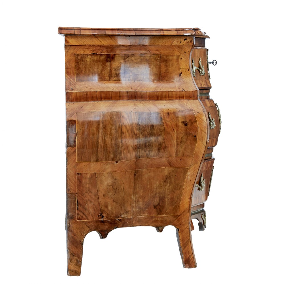 An early nineteenth-century walnut bombe commode featuring three curved drawers with parquetry inlay and ornate brass handles. View VI