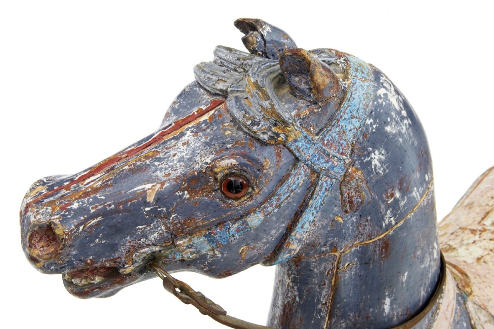 A carved wooden carousel horse sculpture with distressed blue and cream paint, detailed saddle, leather reins, and a raised front leg. View VI
