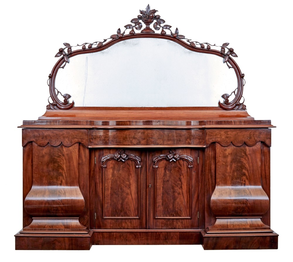 A Victorian flame mahogany sideboard featuring a carved botanical frame around an oval mirror top above cupboard doors and pedestal bases.