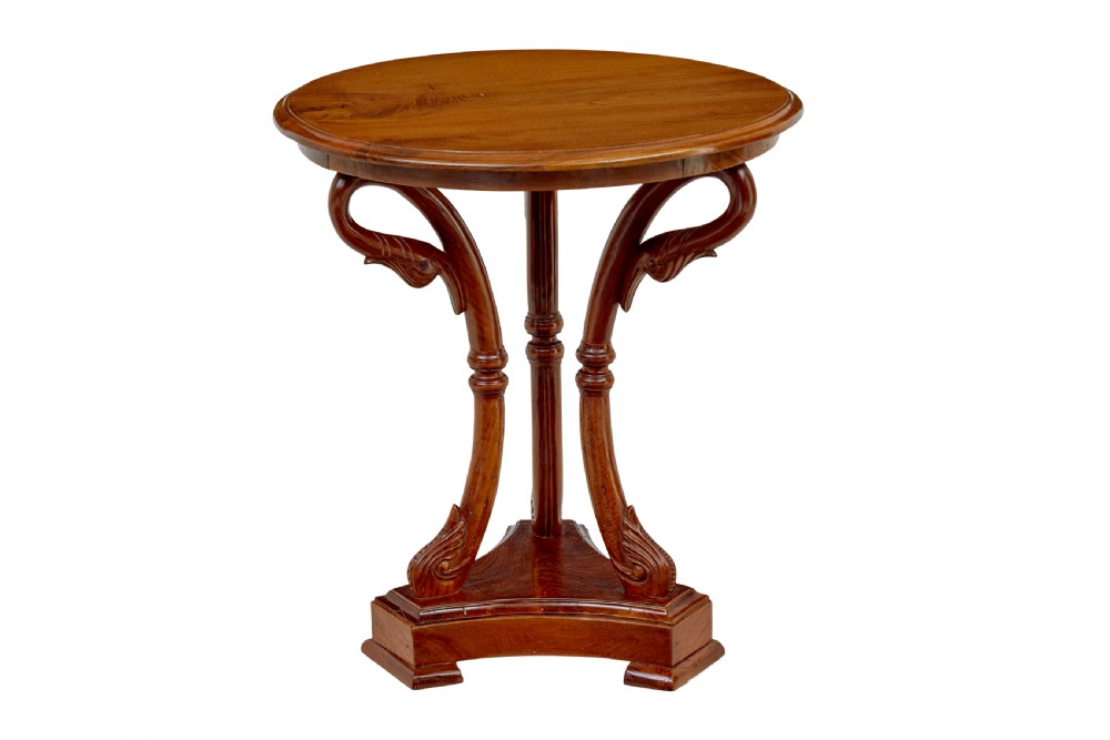 A circular carved teak side table featuring three curved legs and a central turned column on a triangular plinth base.