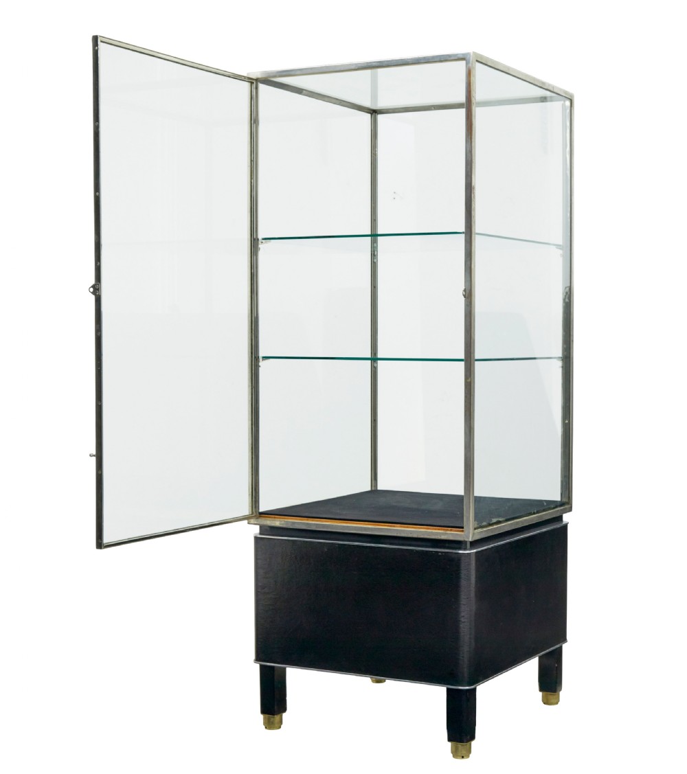 A tall rectangular Art Deco display cabinet featuring transparent glass sides and glass shelves within a thin steel frame, set upon a black leather-clad base with short brass-tipped legs. View II