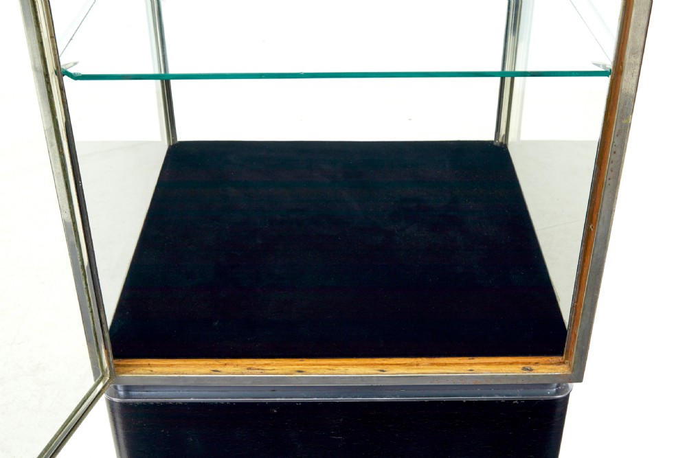 A tall rectangular Art Deco display cabinet featuring transparent glass sides and glass shelves within a thin steel frame, set upon a black leather-clad base with short brass-tipped legs. View VII