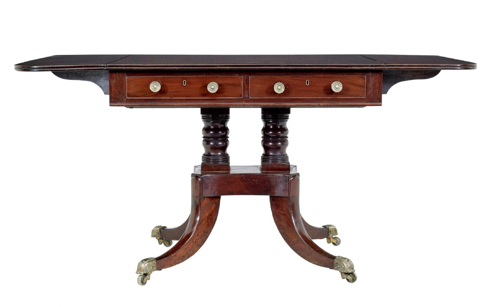 An early Victorian mahogany sofa table featuring two frieze drawers, drop-leaf ends, turned twin column supports, and splayed legs with brass caps and castors.