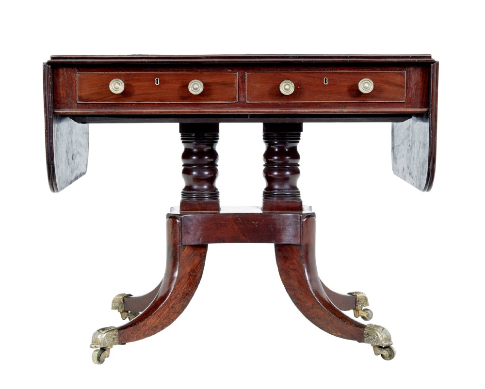 An early Victorian mahogany sofa table featuring two frieze drawers, drop-leaf ends, turned twin column supports, and splayed legs with brass caps and castors. View II