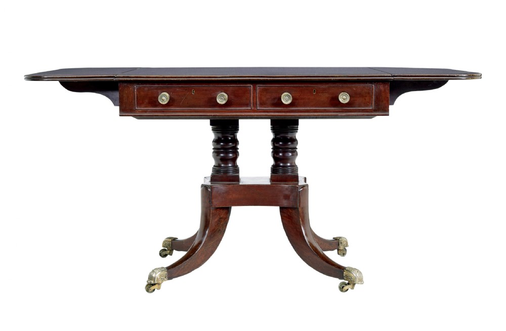 An early Victorian mahogany sofa table featuring two frieze drawers, drop-leaf ends, turned twin column supports, and splayed legs with brass caps and castors. View V