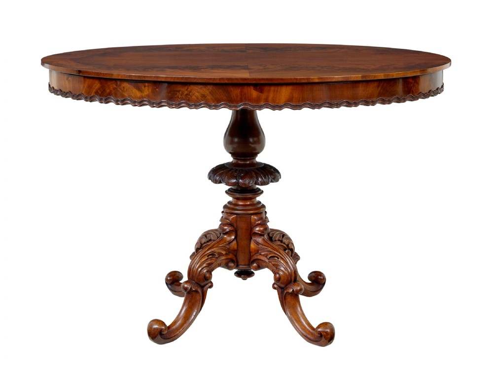 A 19th-century mahogany oval centre table featuring a flame mahogany veneered top, a scalloped apron, and a heavily carved tripod pedestal base with scroll feet.