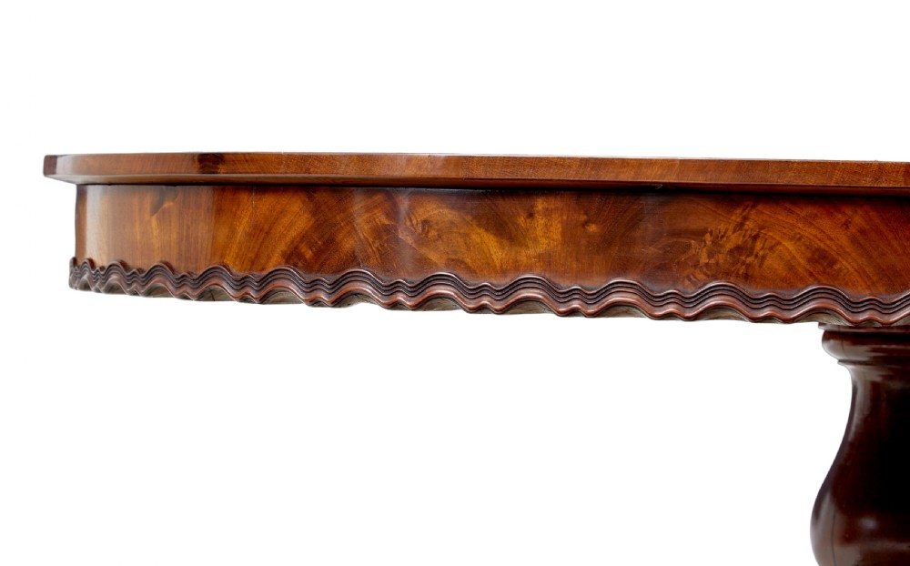 A 19th-century mahogany oval centre table featuring a flame mahogany veneered top, a scalloped apron, and a heavily carved tripod pedestal base with scroll feet. View IV