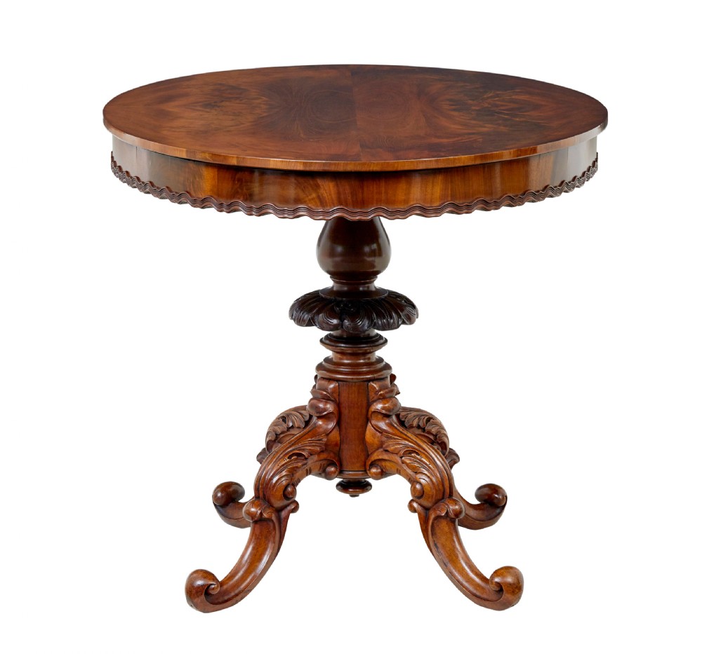 A 19th-century mahogany oval centre table featuring a flame mahogany veneered top, a scalloped apron, and a heavily carved tripod pedestal base with scroll feet. View V