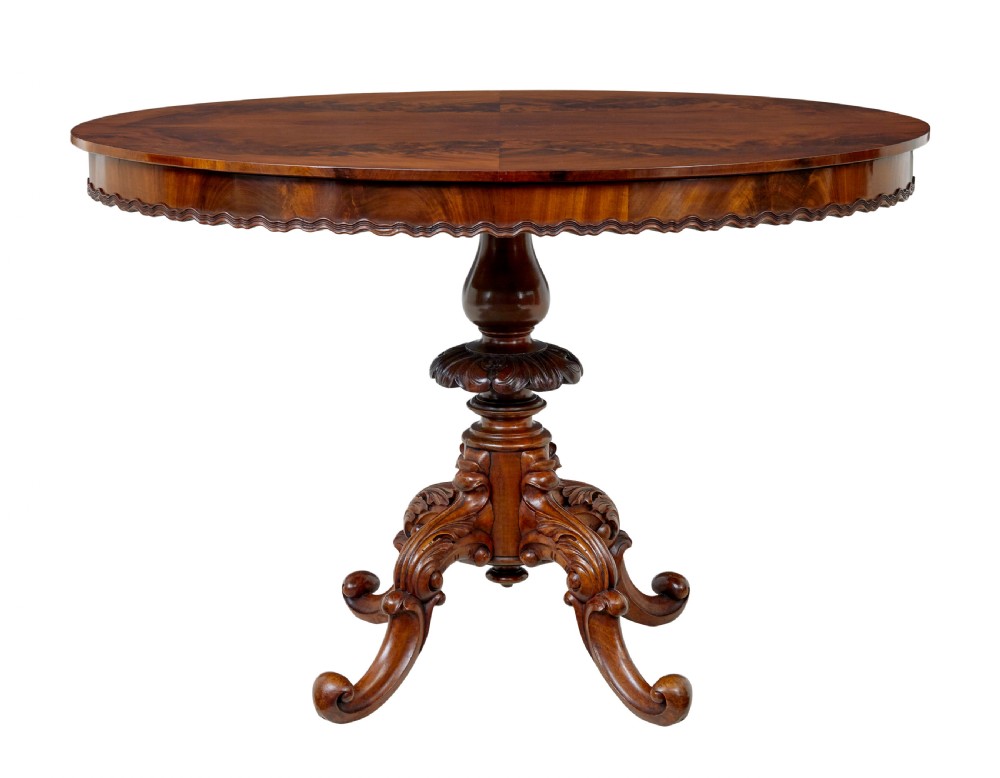 A 19th-century mahogany oval centre table featuring a flame mahogany veneered top, a scalloped apron, and a heavily carved tripod pedestal base with scroll feet. View VI