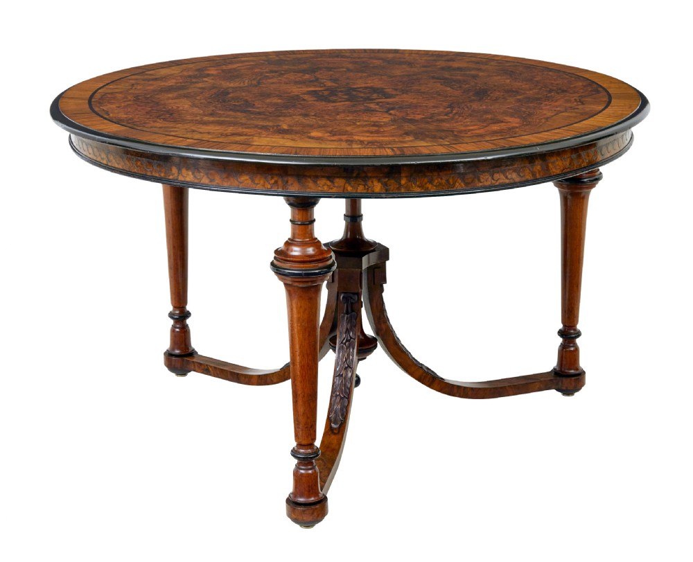 An early 20th-century circular burr walnut occasional table featuring an inlaid top, decorative apron, and three turned legs connected to a central carved triform base.