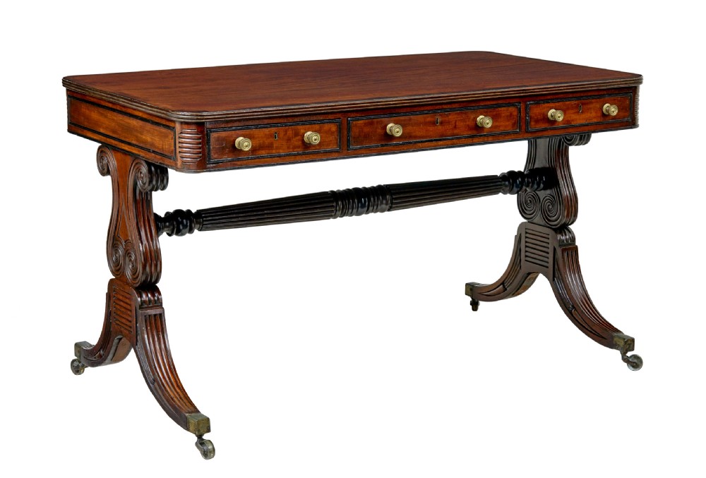 A 19th-century William IV mahogany library desk featuring three frieze drawers with brass knobs, carved scroll end supports, and a turned stretcher on brass castors.