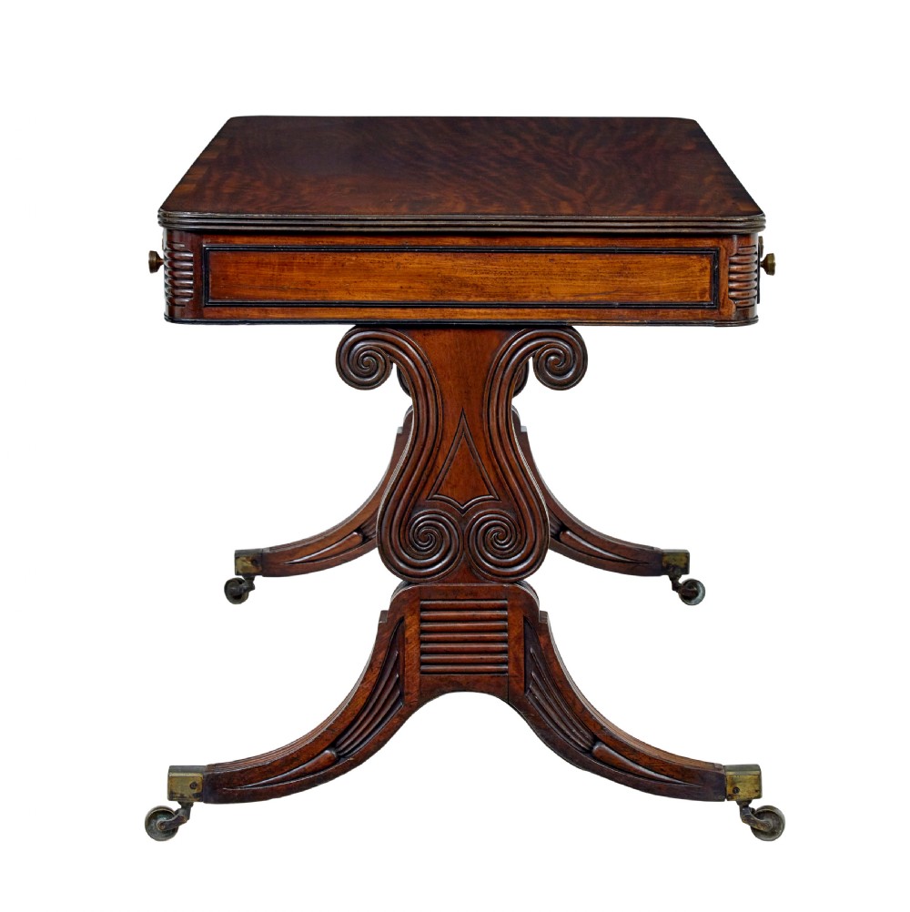 A 19th-century William IV mahogany library desk featuring three frieze drawers with brass knobs, carved scroll end supports, and a turned stretcher on brass castors. View IV