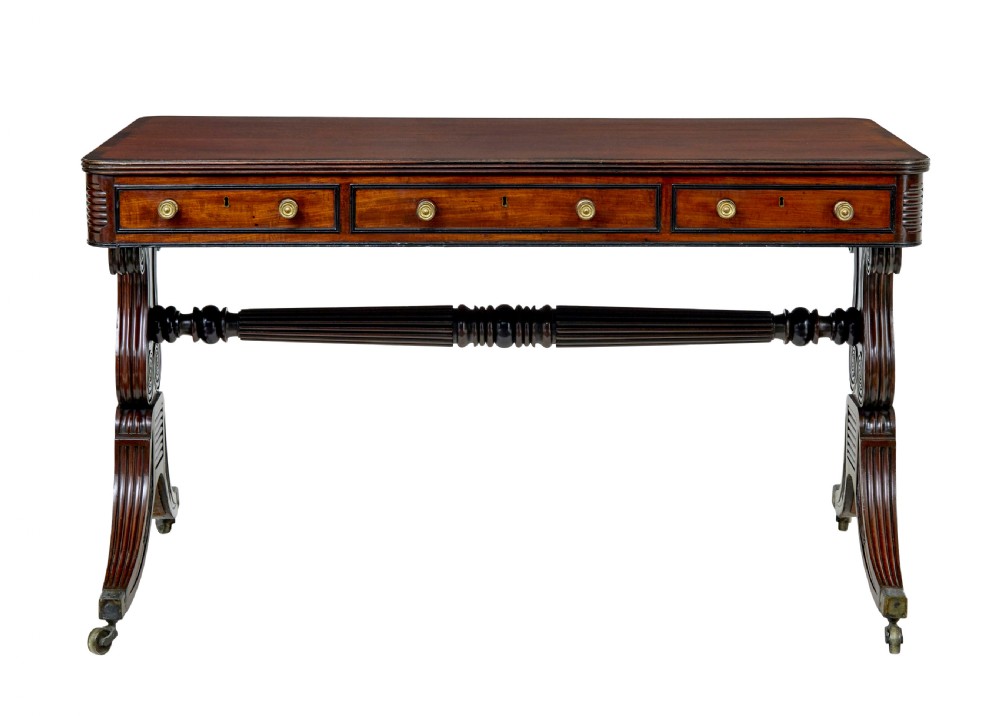 A 19th-century William IV mahogany library desk featuring three frieze drawers with brass knobs, carved scroll end supports, and a turned stretcher on brass castors. View V