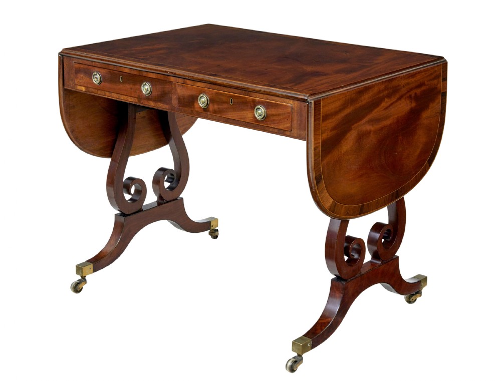 An early 19th-century mahogany sofa table featuring two drop leaves, twin frieze drawers with brass handles, and lyre-shaped scroll legs on brass castors.
