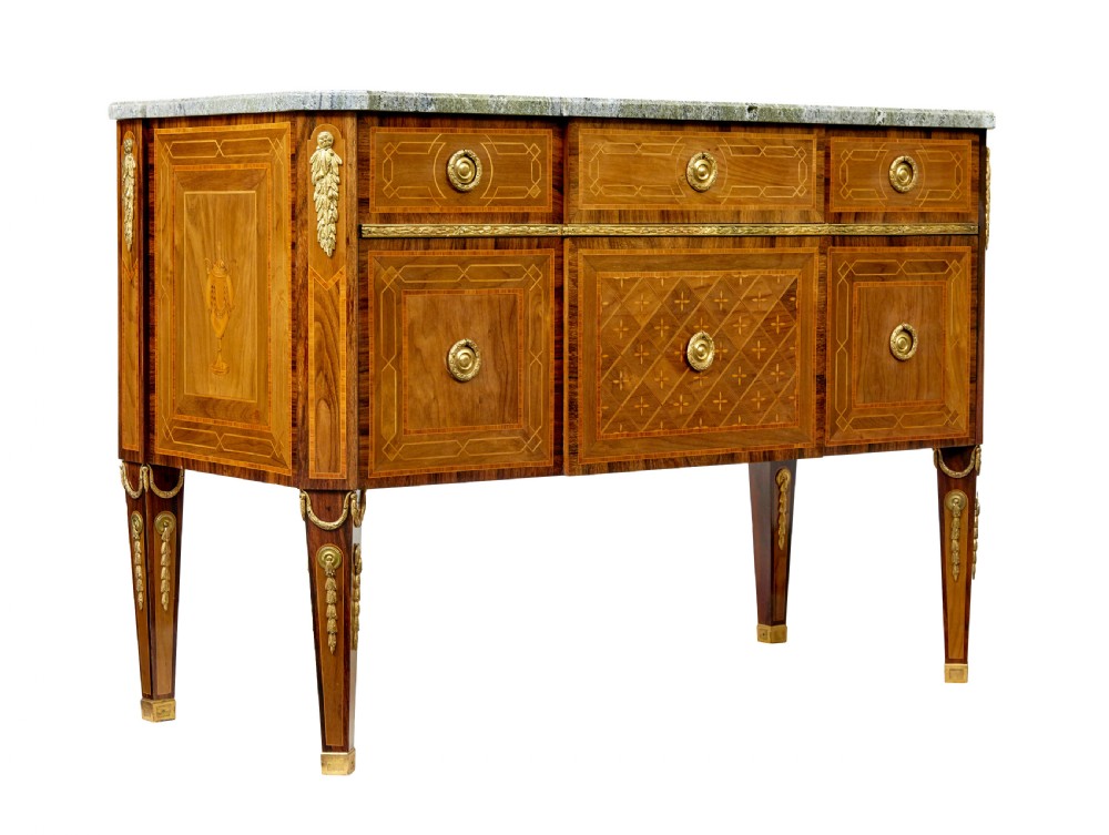 An early 20th-century walnut commode featuring marquetry inlay, gilt bronze mounts, square tapered legs, and a fitted grey marble top.