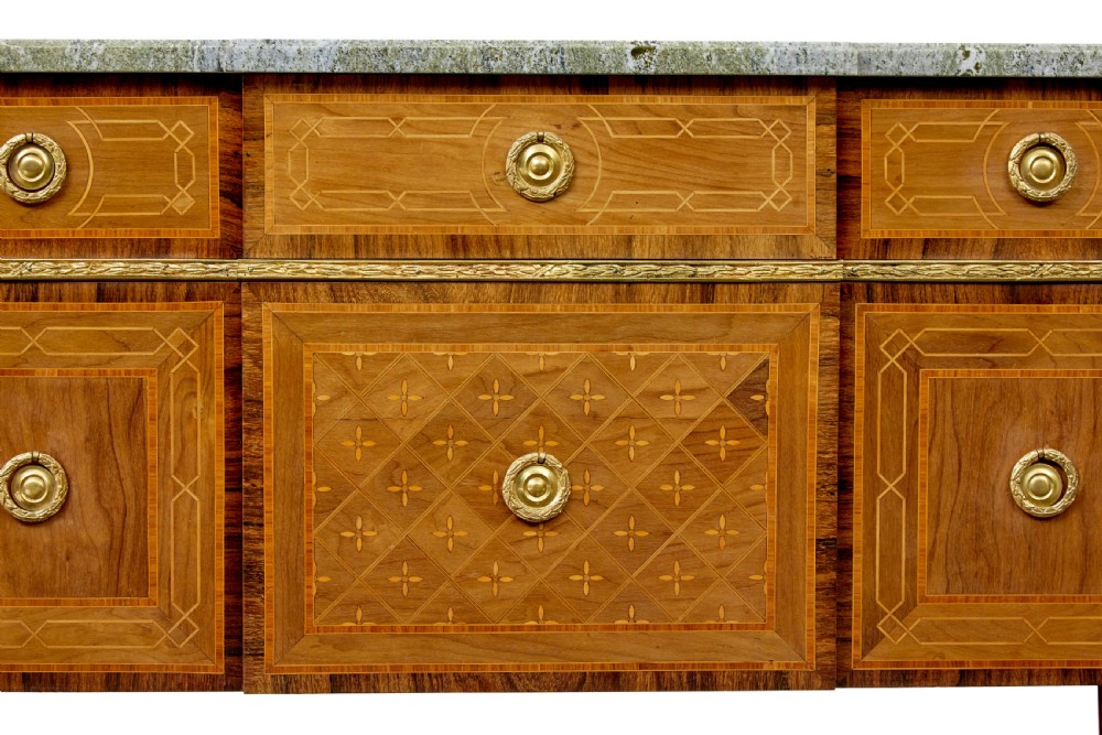 An early 20th-century walnut commode featuring marquetry inlay, gilt bronze mounts, square tapered legs, and a fitted grey marble top. View IX