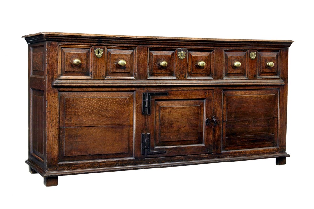 An 18th-century dark oak dresser featuring three geometric panelled drawers with brass knobs above cupboard doors with iron hinges.