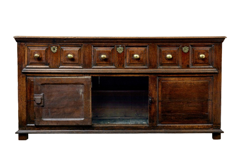 An 18th-century dark oak dresser featuring three geometric panelled drawers with brass knobs above cupboard doors with iron hinges. View V