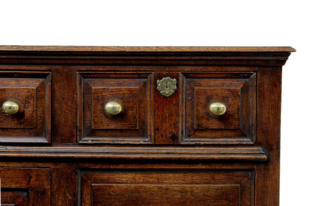 An 18th-century dark oak dresser featuring three geometric panelled drawers with brass knobs above cupboard doors with iron hinges. View X
