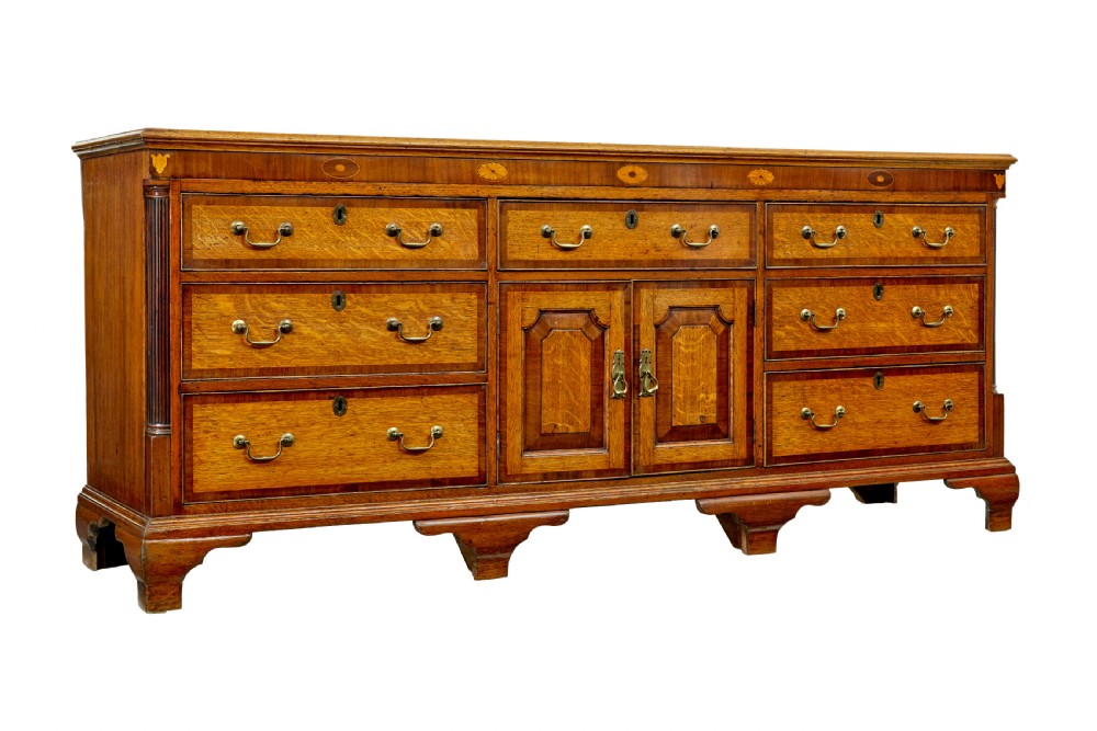 An 18th-century inlaid oak Lancashire dresser base featuring six drawers, a central two-door fielded panel cupboard, brass handles, and ogee bracket feet.