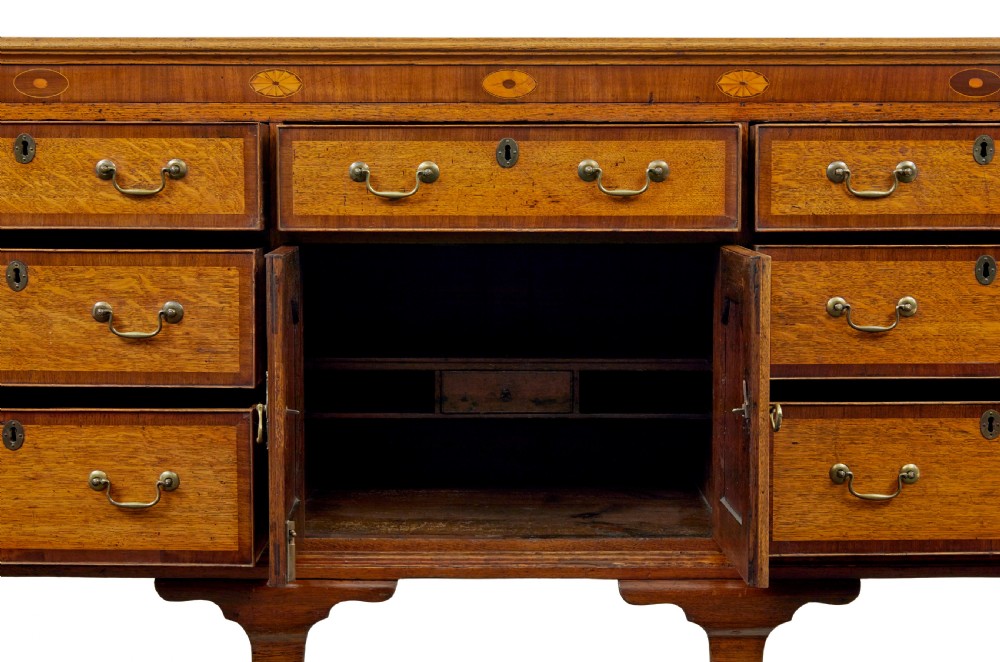 An 18th-century inlaid oak Lancashire dresser base featuring six drawers, a central two-door fielded panel cupboard, brass handles, and ogee bracket feet. View V
