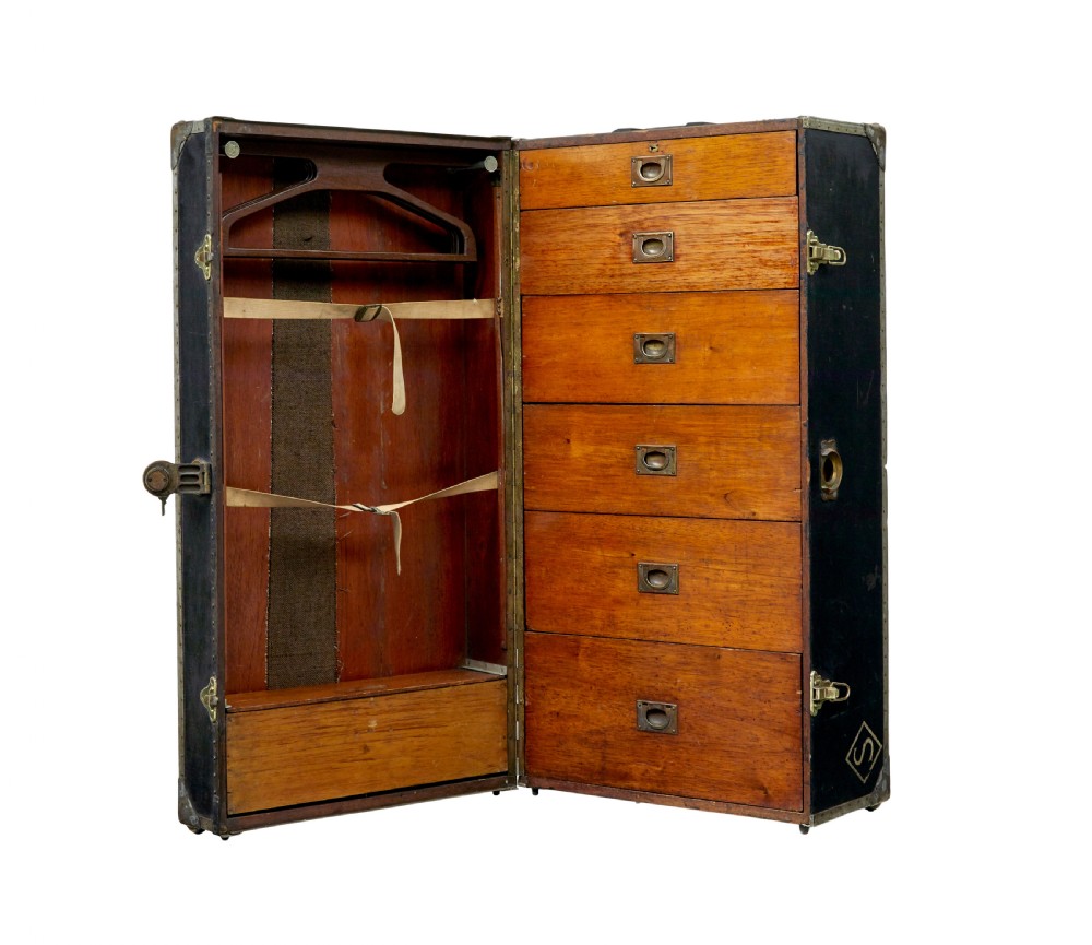 An open early 20th-century vertical steamer chest featuring a clothes hanging compartment with straps on the left and a column of six wooden drawers with recessed brass handles on the right.