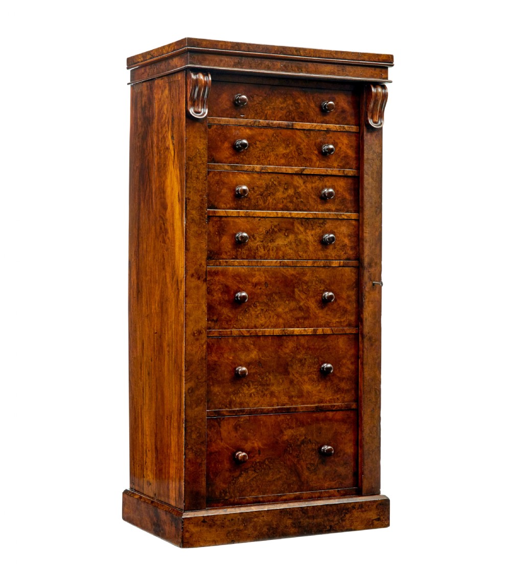 A tall, narrow Victorian burr walnut Wellington chest featuring seven graduated drawers with turned knob handles, decorative scroll brackets, and a locking side pilaster set upon a plinth base.