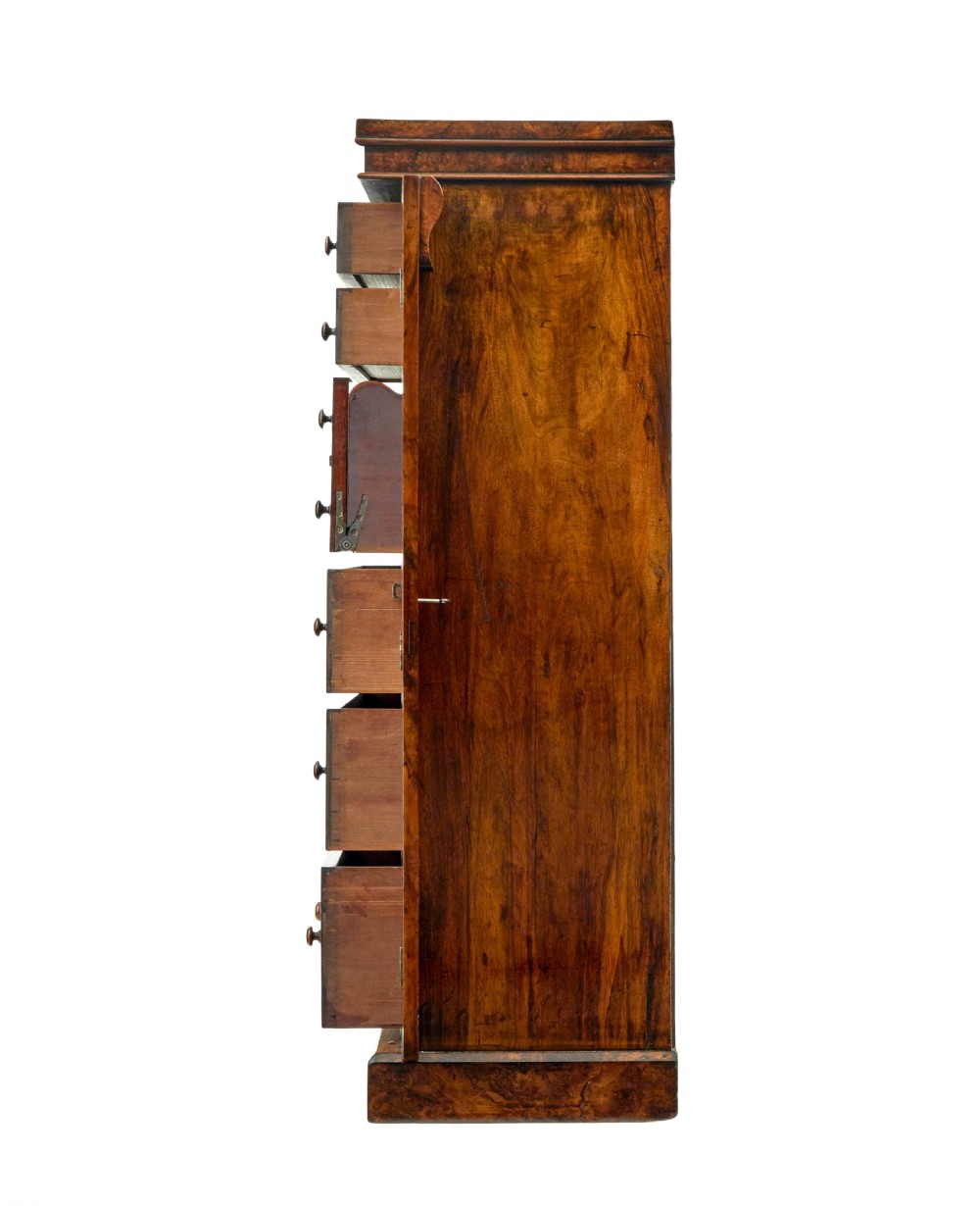 A tall, narrow Victorian burr walnut Wellington chest featuring seven graduated drawers with turned knob handles, decorative scroll brackets, and a locking side pilaster set upon a plinth base. View V